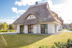 Ein reetgedecktes Haus mit weißer Fassade, großen Fenstern und einem gepflegten Rasen.