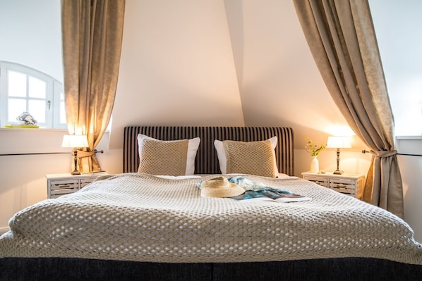 Un dormitorio con una cama grande, cabecera a rayas, dos mesitas de noche con lámparas y una ventana con cortinas.