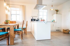 A modern kitchen with a central island, wooden dining table with blue cushions, and pendant lighting.