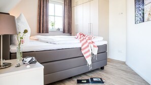 A bedroom with a bed, a bedside table, a lamp, a window with curtains, and a wall with framed pictures.
