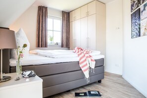A bedroom with a bed, a bedside table, a lamp, a window with curtains, and a wall with framed pictures.