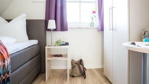 Una camera da letto con un letto, un comodino con una lampada, una piccola mensola con dei libri, una borsa e una finestra con tende.