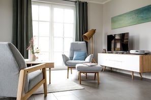 A modern living room with a grey sofa, a matching armchair, a wooden coffee table, a side table with a vase, a floor lamp, a TV, and a white cabinet.
