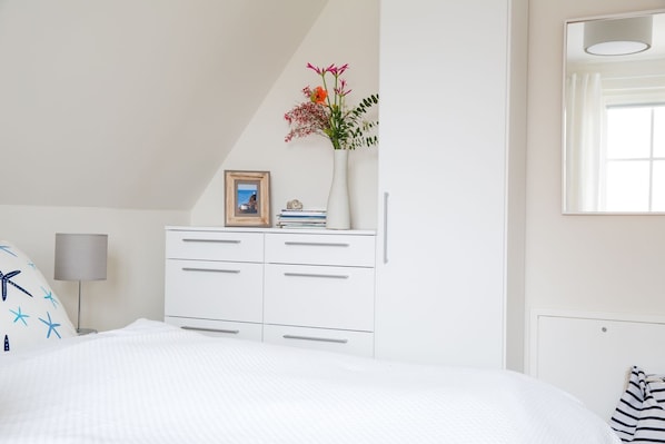 A bedroom with a white bed, a dresser with drawers, a framed photo, and a vase with flowers.