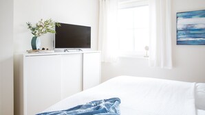 Une chambre à coucher avec un lit, un téléviseur posé sur une commode blanche, une fenêtre avec des rideaux blancs et un tableau accroché au mur.