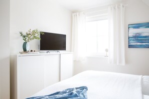 Een slaapkamer met een bed, een televisie op een witte dressoir, een raam met witte gordijnen, en een schilderij aan de muur.