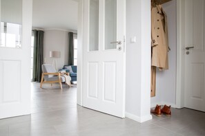 A modern living room with a sofa and a chair, a coat rack with a coat and shoes, and a door leading to another room. - Vacation home Seeschwalbe 33 - Holiday home Seeschwalbe 33 - Close to nature (Fuhlendorf)