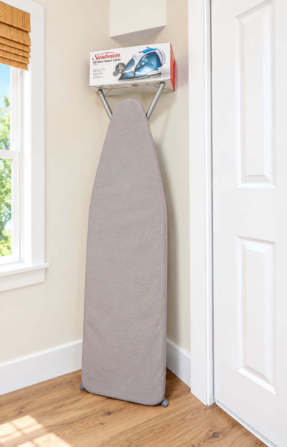 Une planche à repasser grise, sur laquelle repose une boîte de la marque Sunbeam, est placée contre une porte et un mur blancs. On aperçoit une fenêtre et un plancher en bois.