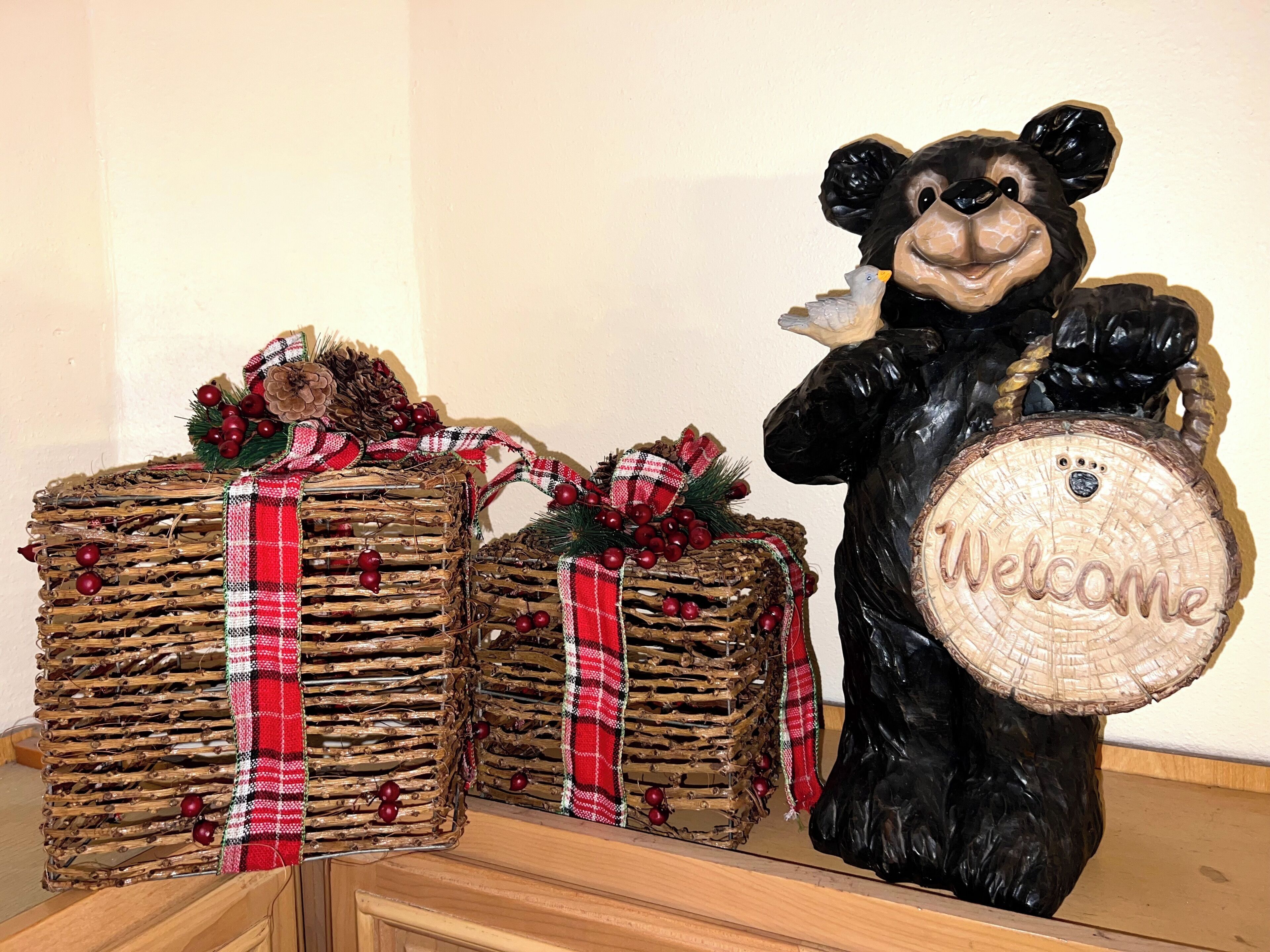 Un orso decorativo che regge un cartello di benvenuto, accanto a due composizioni simili a pacchetti regalo con fiocchi rossi e bianchi.