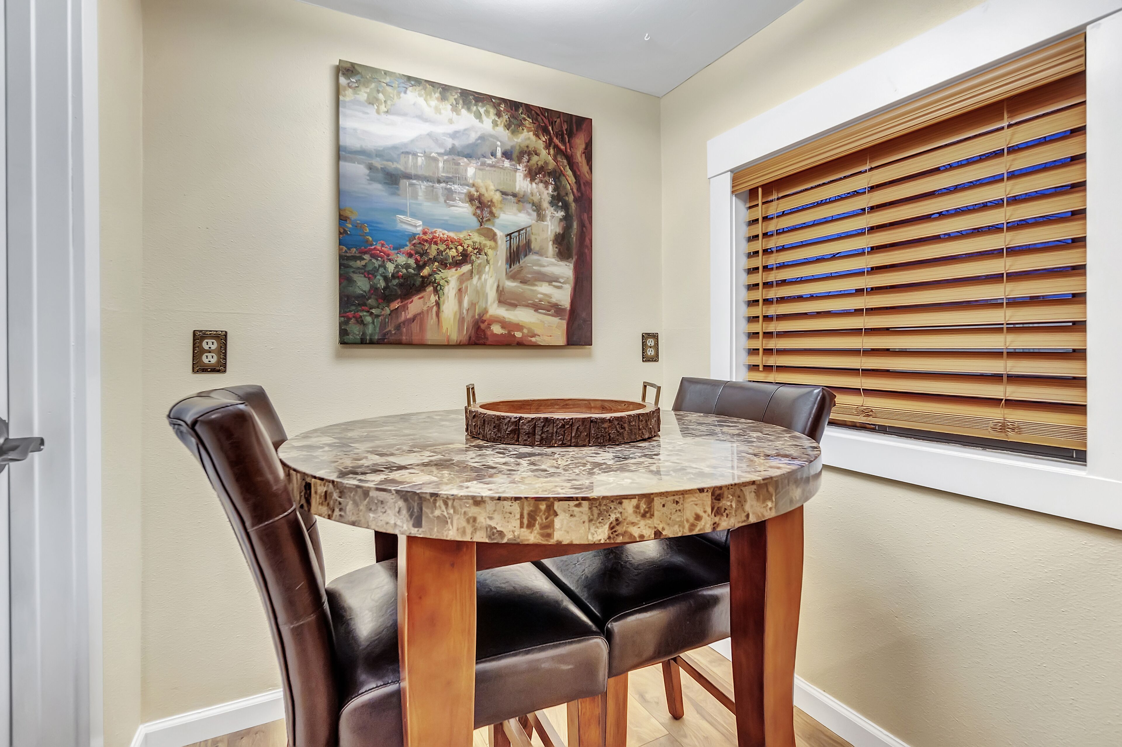 Un coin repas avec une table ronde, des chaises en cuir noir, un tableau au mur et des stores en bois à la fenêtre.