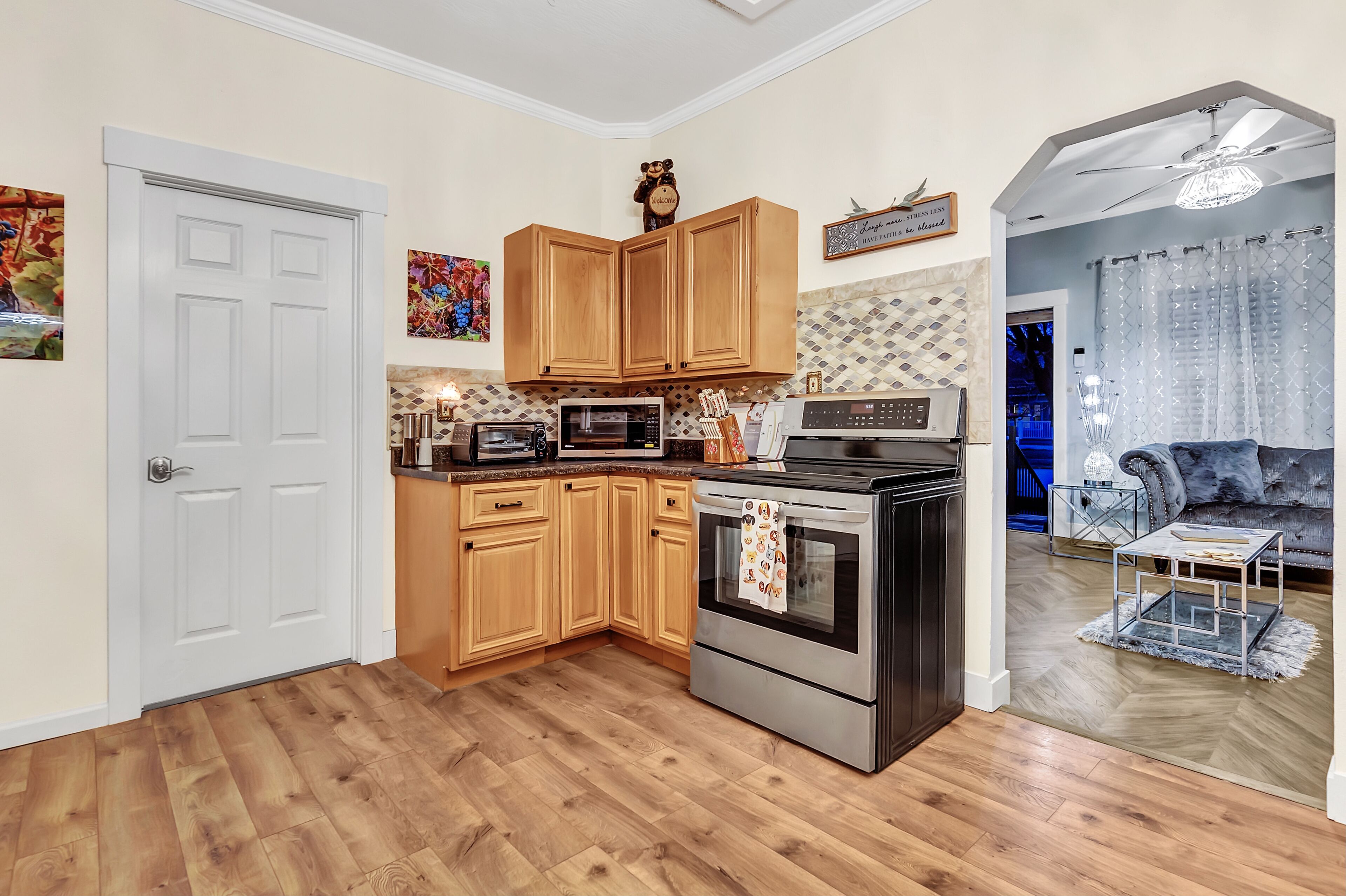 Een moderne keuken met houten kastjes, een roestvrijstalen oven en een magnetron. Er is een deur naar een andere kamer en een zithoek met een blauwe bank.