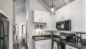 A modern kitchen with white cabinets, a black countertop, stainless steel appliances, and a hanging light fixture. - Heavensent Tiny Home @ The Retreat at Water's Edge - Lake Views! (Tracy City)