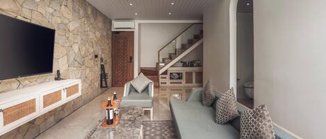 A modern living room with a stone wall, a flat-screen TV, a marble coffee table, a light blue sofa with patterned cushions, and a staircase leading to another level.