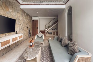 Una sala de estar moderna con una pared de piedra, un televisor de pantalla plana, una mesa de centro de mármol, un sofá azul claro con cojines estampados y una escalera que sube a otro nivel.