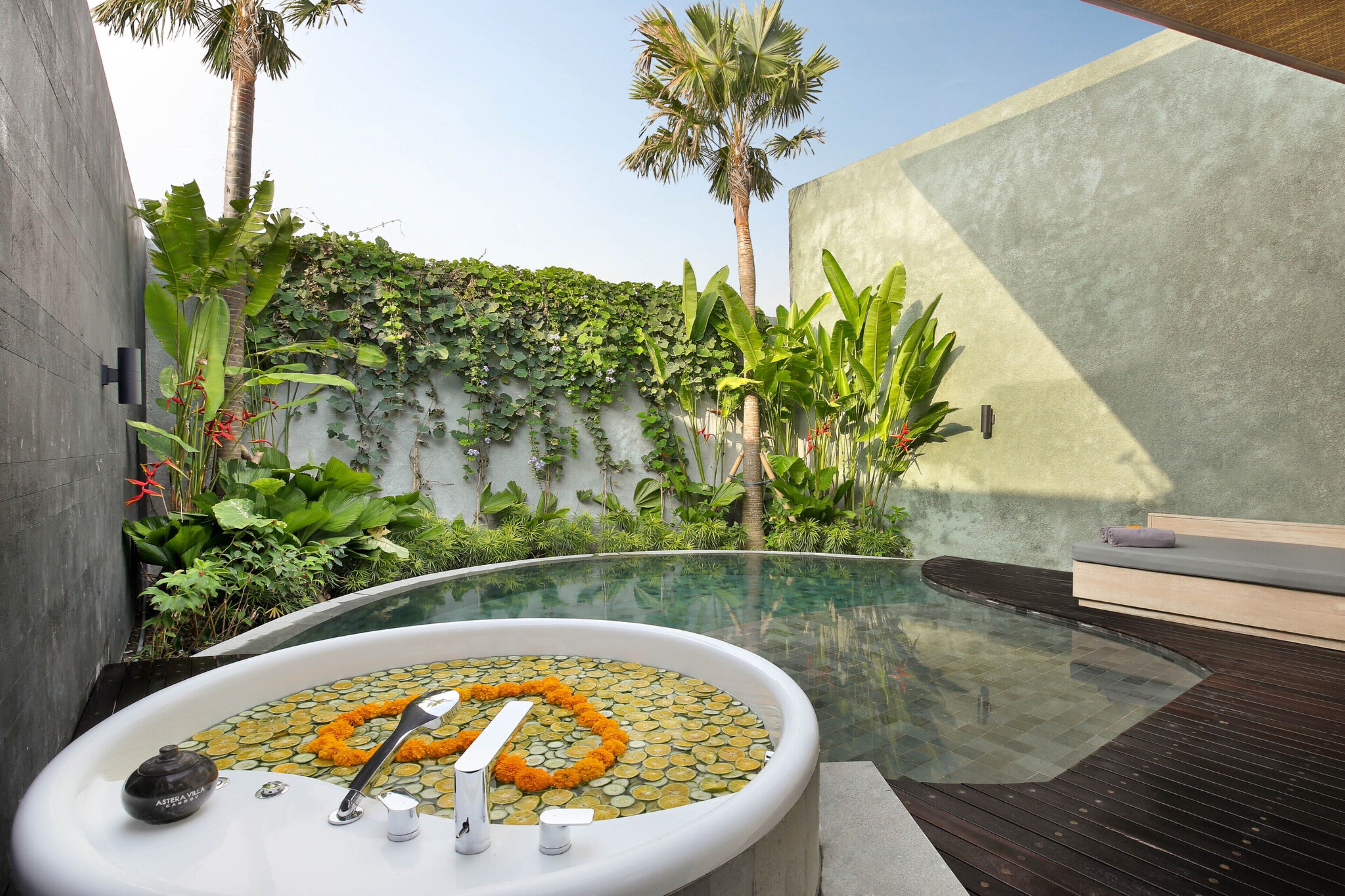 A modern outdoor hot tub with a stone mosaic design, surrounded by lush greenery and a concrete wall.