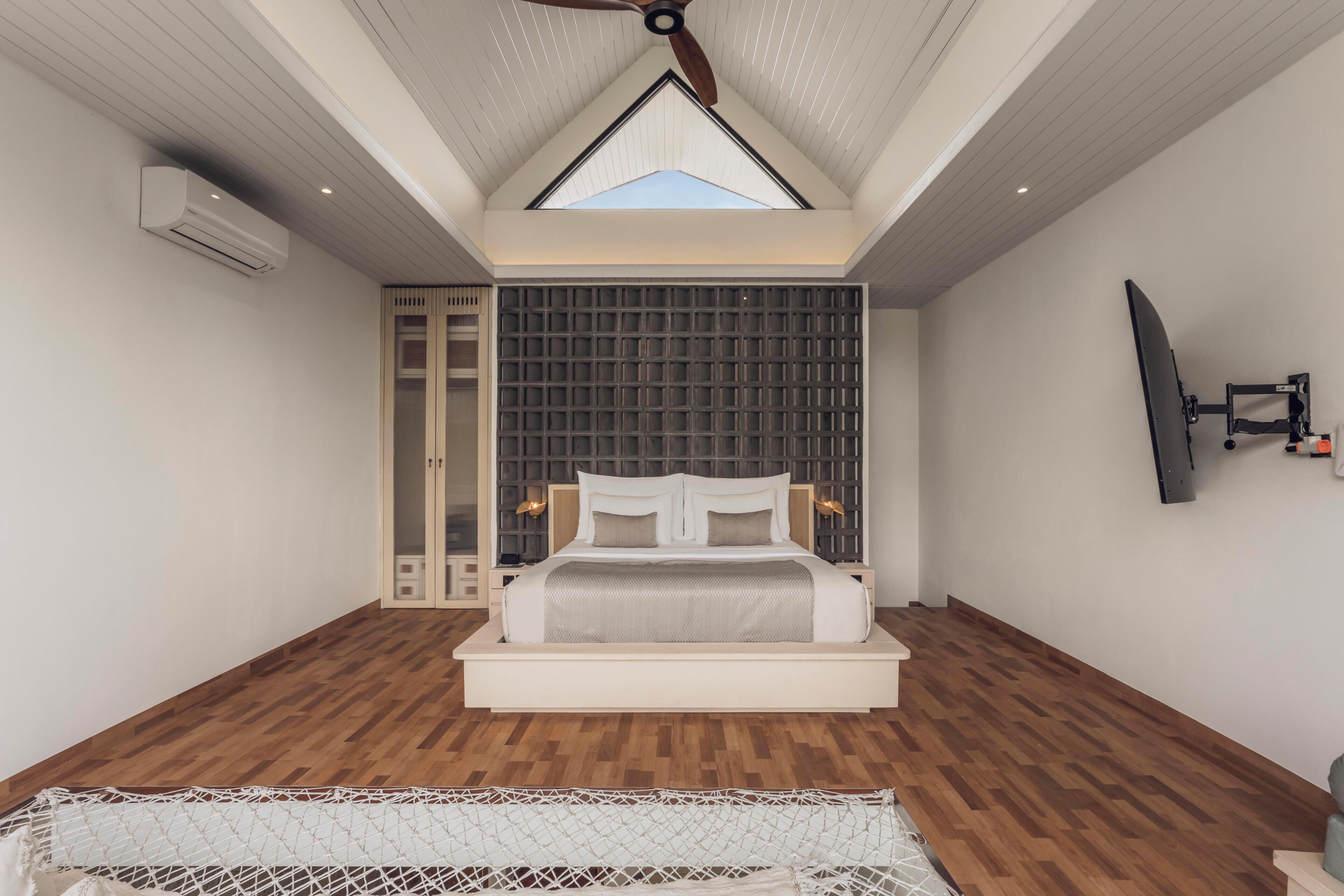 A bedroom with a sloped ceiling, a bed, a TV mounted on the wall, and a window.