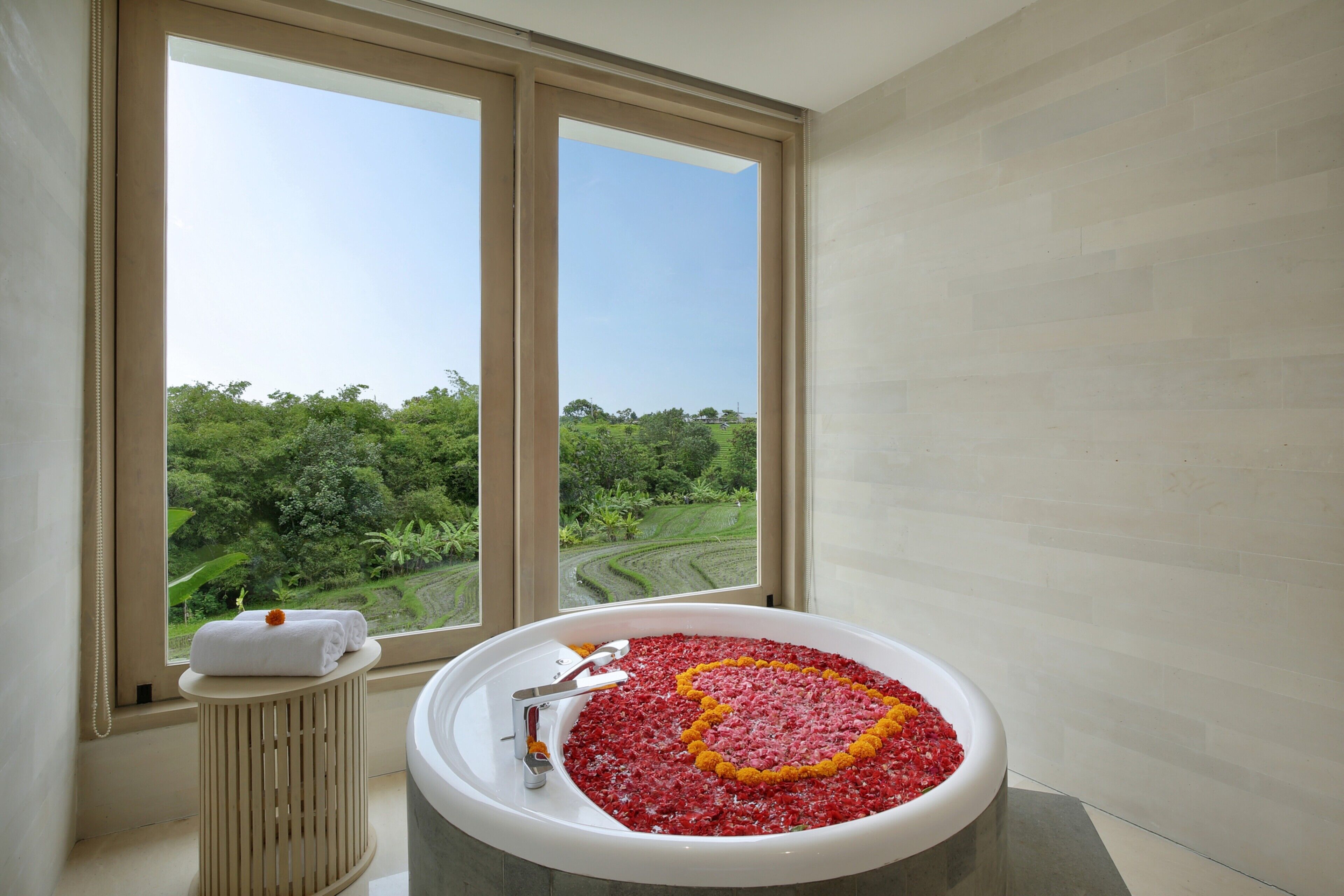 Ein Badezimmer mit einer mit Rosenblüten gefüllten Badewanne, einem Handtuch auf einem Beistelltisch und Blick auf eine üppige Landschaft durch große Fenster.