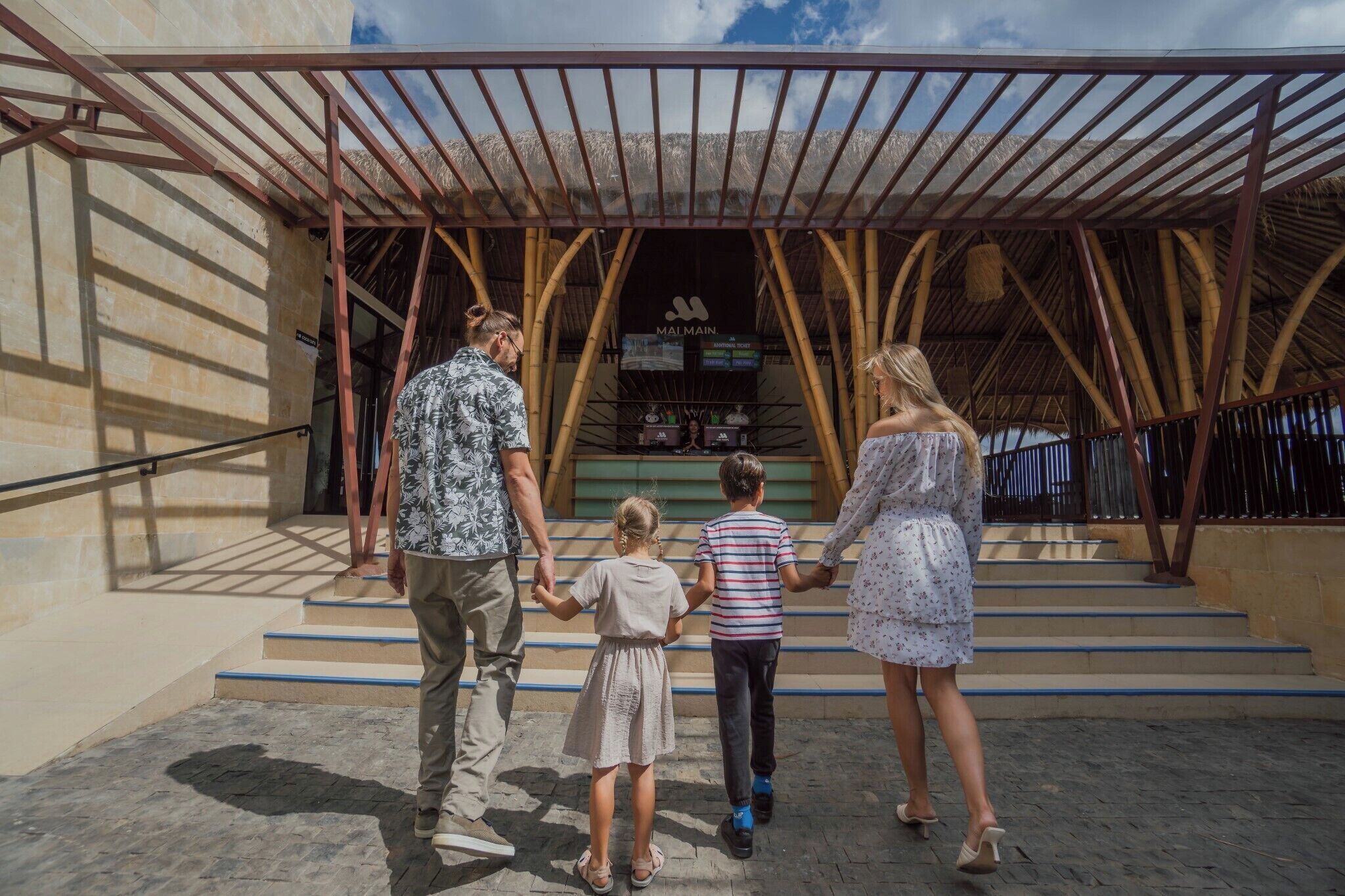 A family walking towards a building with a bamboo structure and a sign that reads 'MALMAIN'.