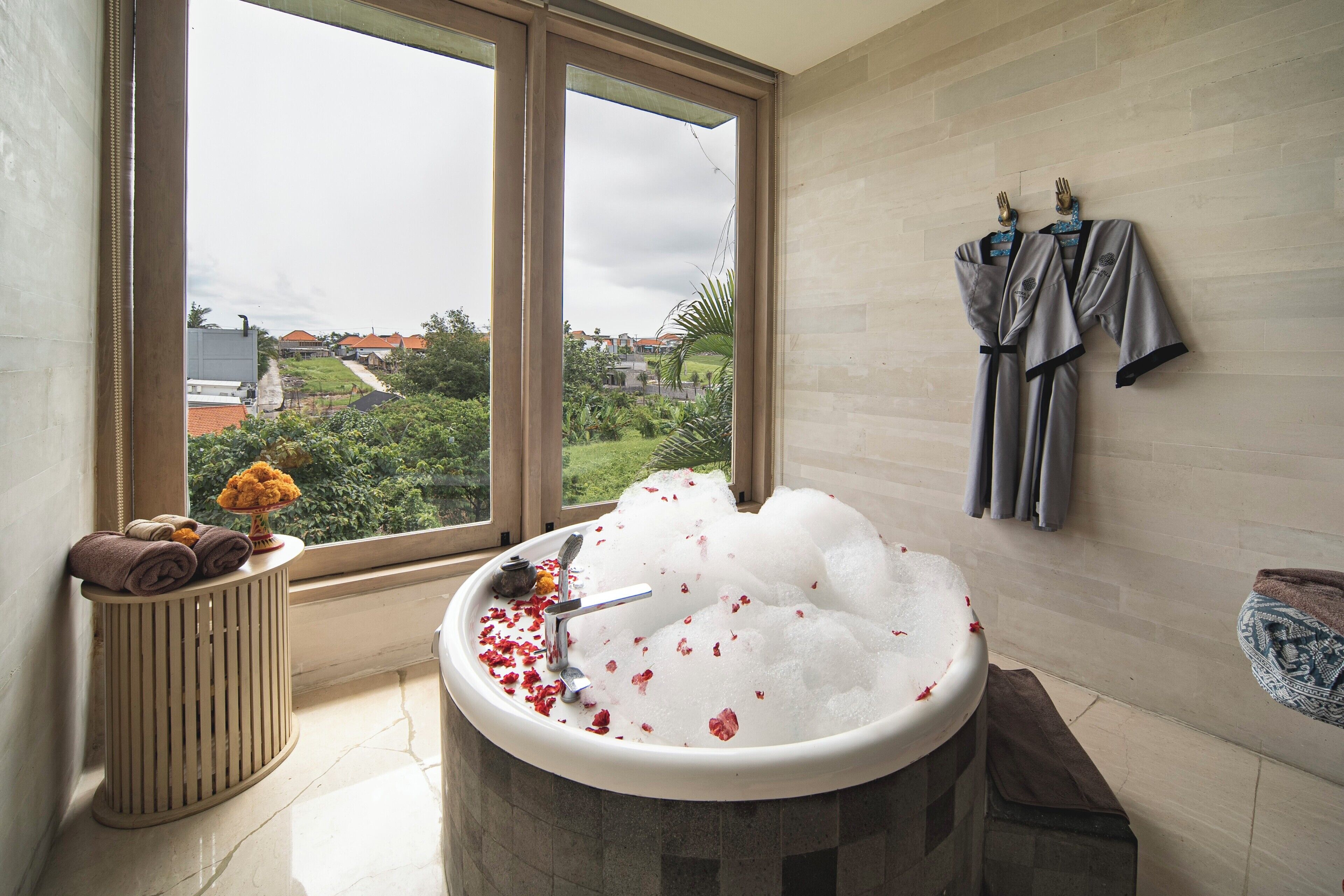 A bathroom with a large window, a bathtub filled with bubbles, towels, and robes hanging on the wall.