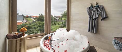 A bathroom with a large window, a bathtub filled with bubbles, towels, and robes hanging on the wall.