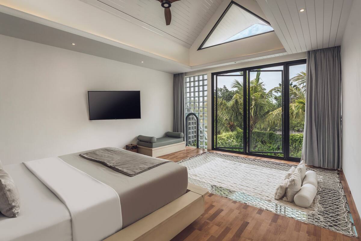 Ein modernes Schlafzimmer mit Bett, Fernseher, Balkon und Blick auf Grünflächen, sowie einem Deckenventilator.