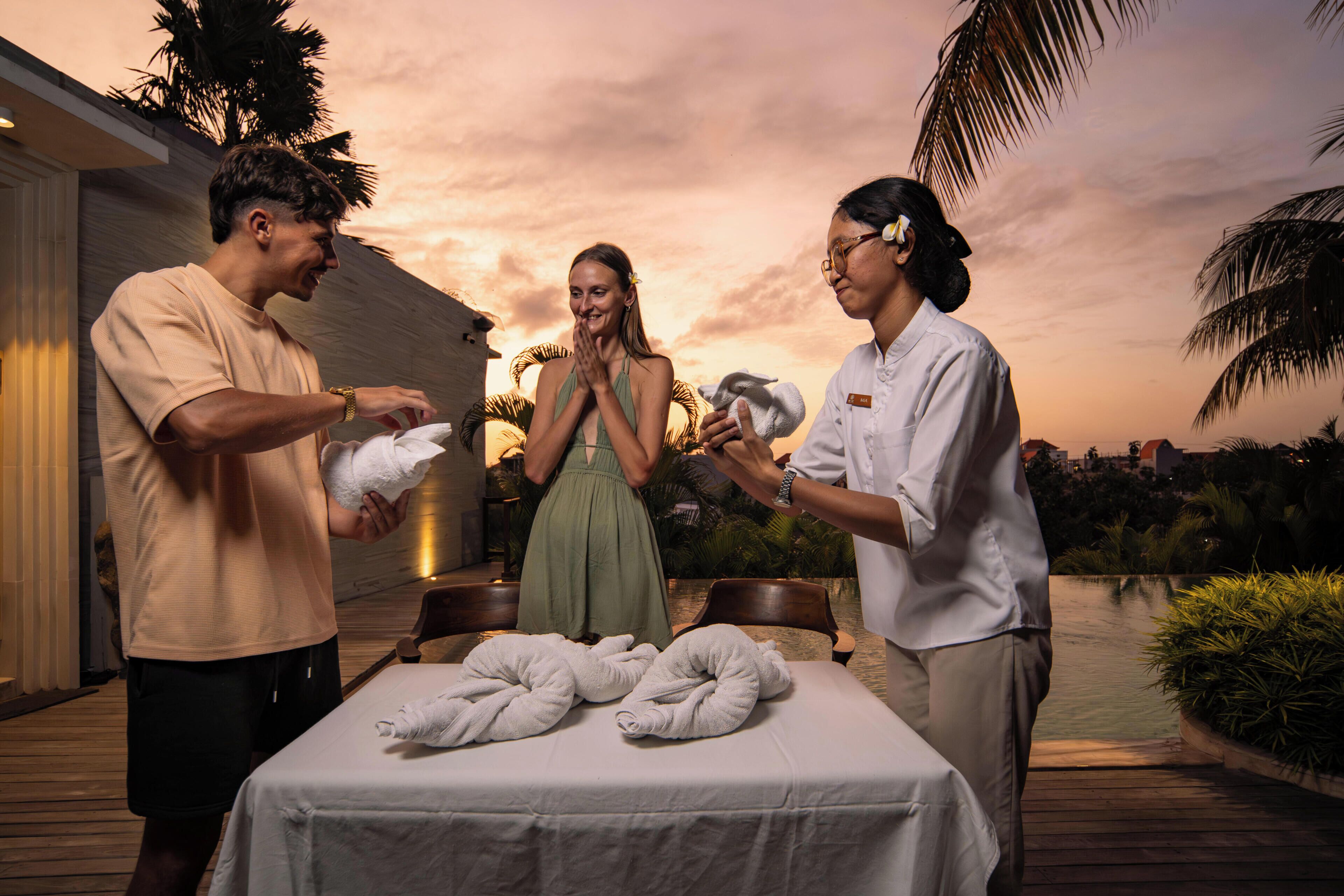 A couple on a balcony with a sunset view, a table with towels, and a pool.