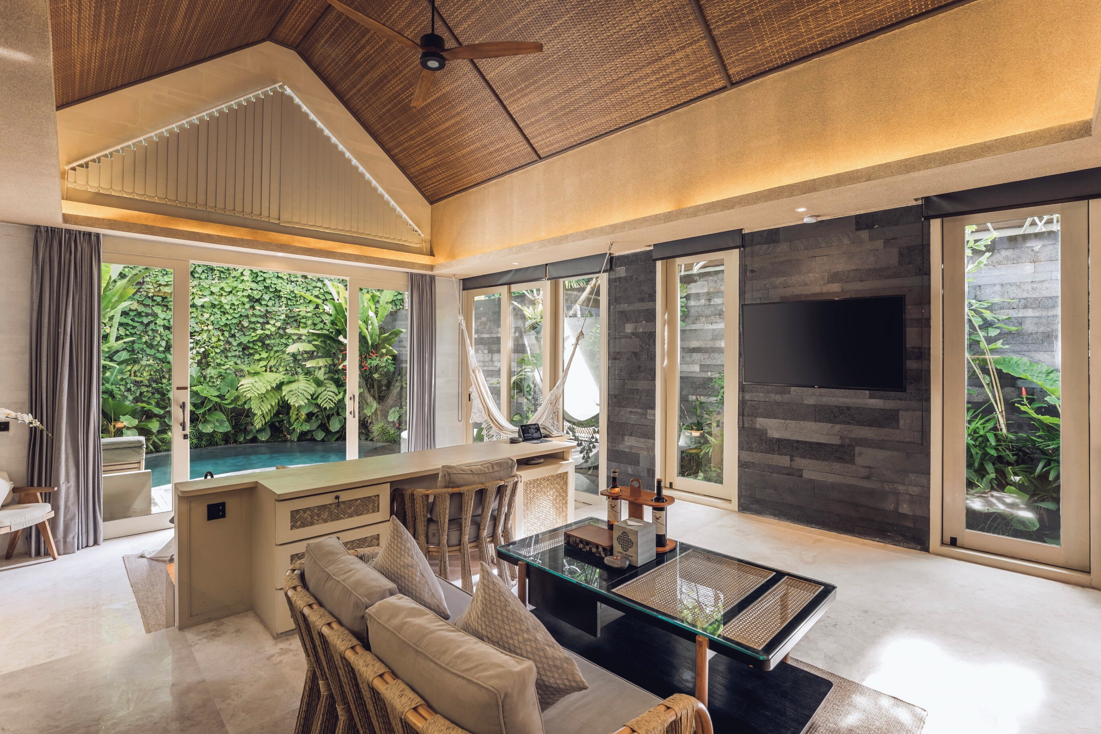 A modern living room with a glass coffee table, wicker sofa, and a TV mounted on a stone wall.