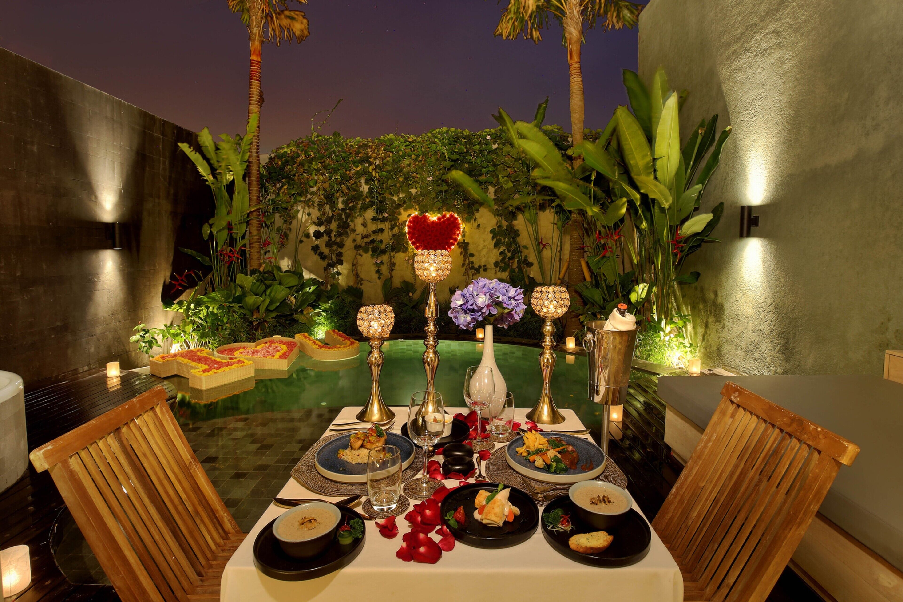 An outdoor dining setup with a table set for two, surrounded by plants and decorative lighting.