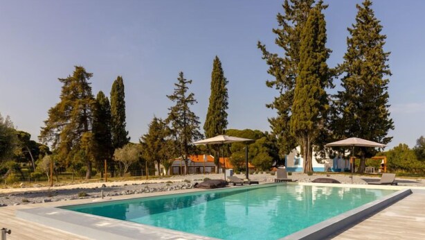 A rectangular swimming pool with a wooden deck surrounding it, two umbrellas, and tall trees in the background. - Quinta de Santa Emilia 1 by Interhome (Alcácer do Sal)