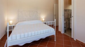 A white metal bed with a decorative headboard in a room with tiled flooring and a door leading to a bathroom.