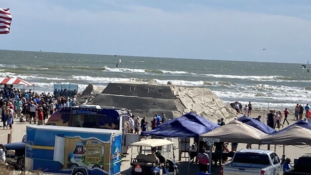 Un espace de stationnement situé au bord de la plage, avec un camion-restaurant, des tentes et une grande formation rocheuse ornée d’inscriptions.
