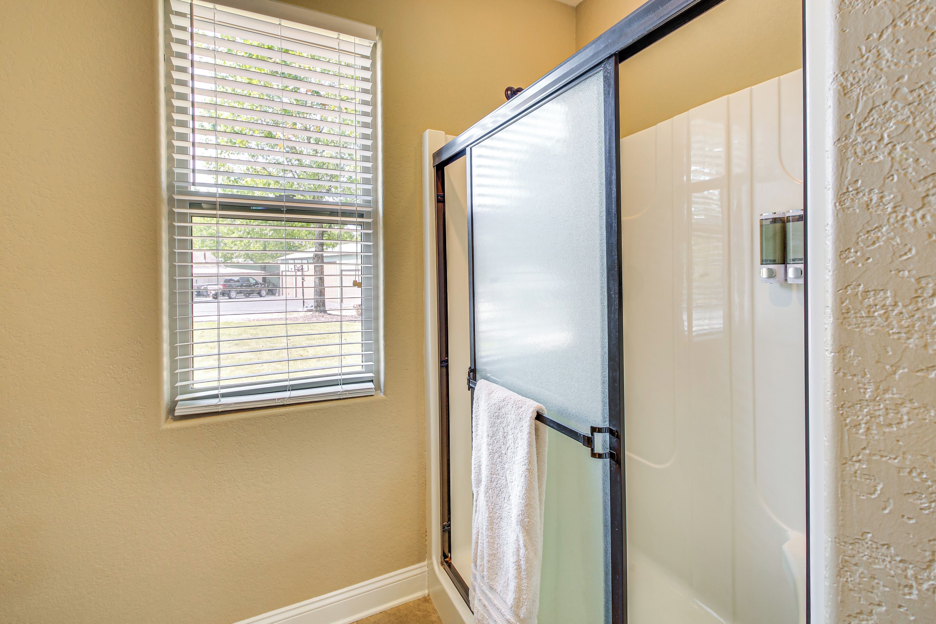 A bathroom with a glass shower door, a window with blinds, and a towel hanging on the door.