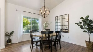 Un coin repas comprenant une table ronde, six chaises, un lustre, une grande fenêtre avec des rideaux et une plante en pot.