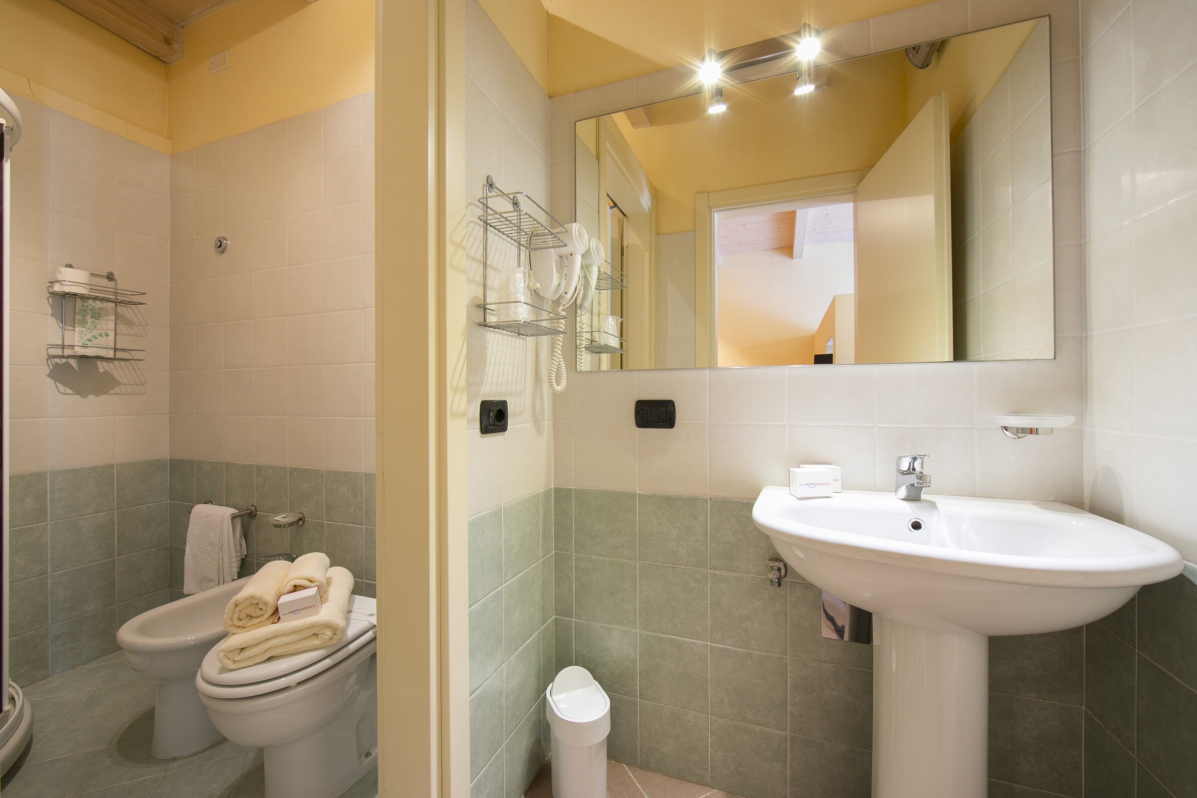 Un baño con inodoro, bidé y lavabo. Hay toallas en el inodoro, un cubo de basura y un espejo sobre el lavabo.