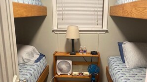 Ein Schlafsaalzimmer mit Etagenbetten, einem Schreibtisch mit Lampe, einem Ventilator und einem Fenster mit Jalousien.