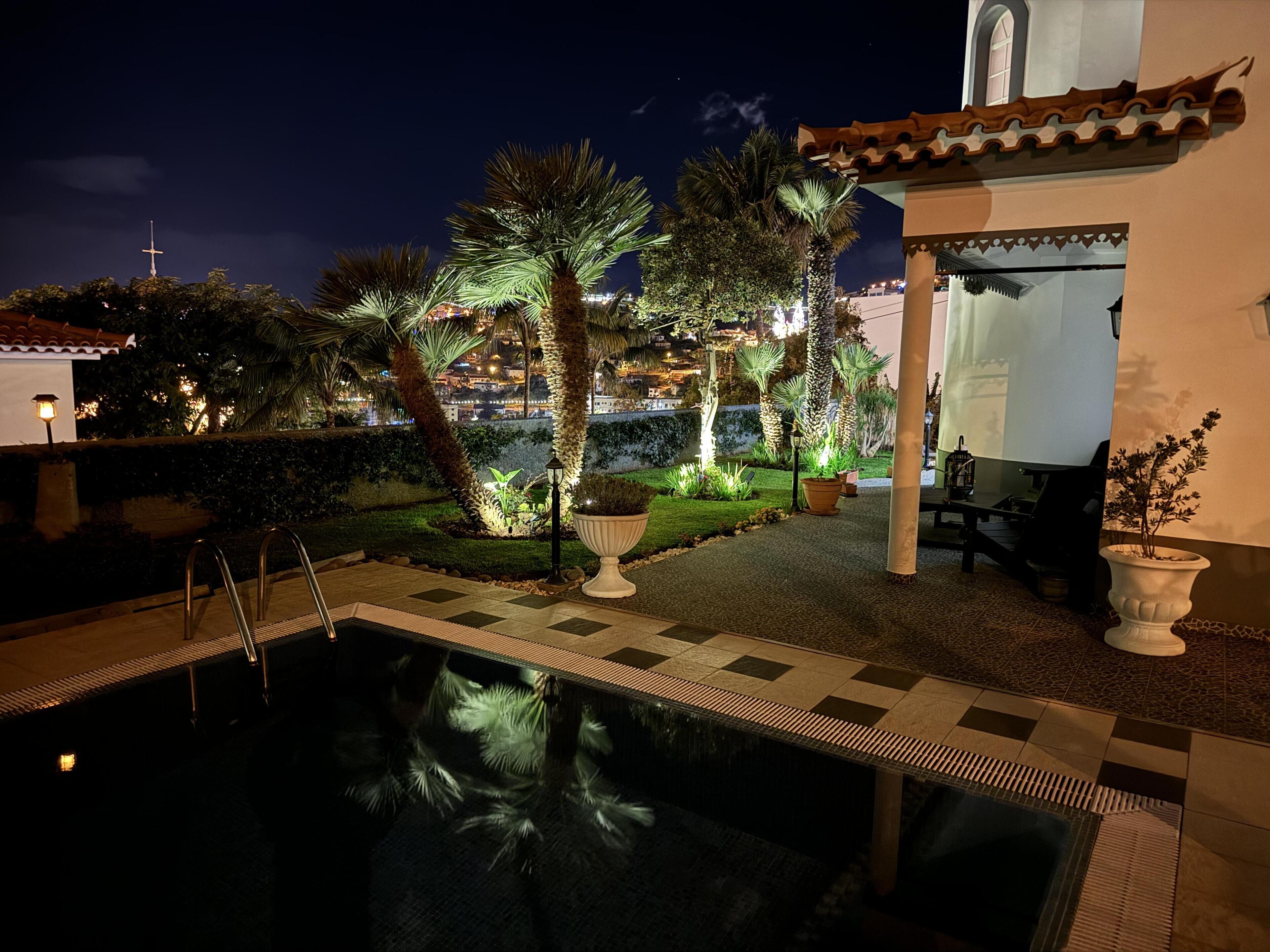 A nighttime view of a swimming pool area with a tiled deck, palm trees, and a building with a cross on top.