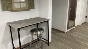 Una habitación con una mesa, un taburete, una ventana con contraventanas y una puerta con espejo.