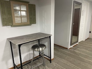 Una habitación con una mesa, un taburete, una ventana con contraventanas y una puerta con espejo.