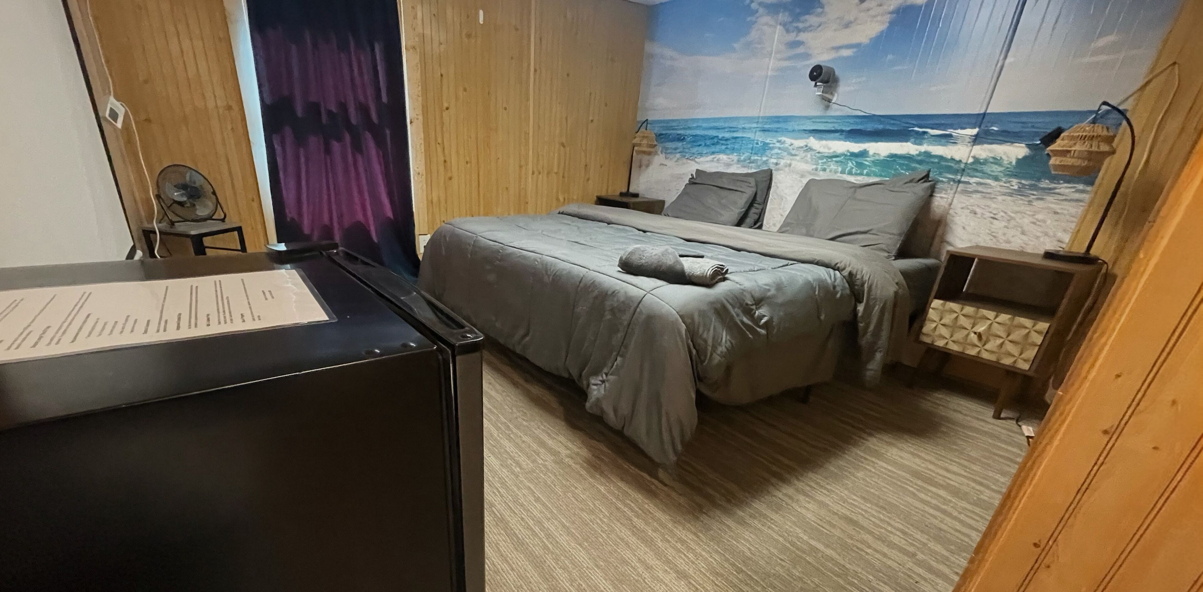 Un dormitorio con cama, escritorio, ventilador y un mural en la pared con vista al océano.