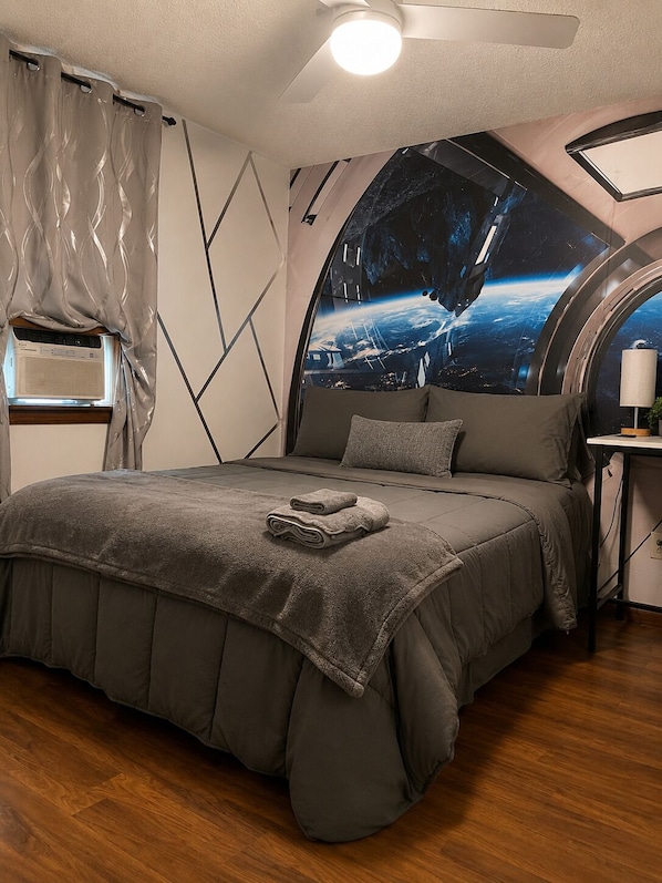 Un dormitorio con techo curvo, un ventanal con vistas a la Tierra, una cama con edredón gris y una mesita de noche con lámpara.