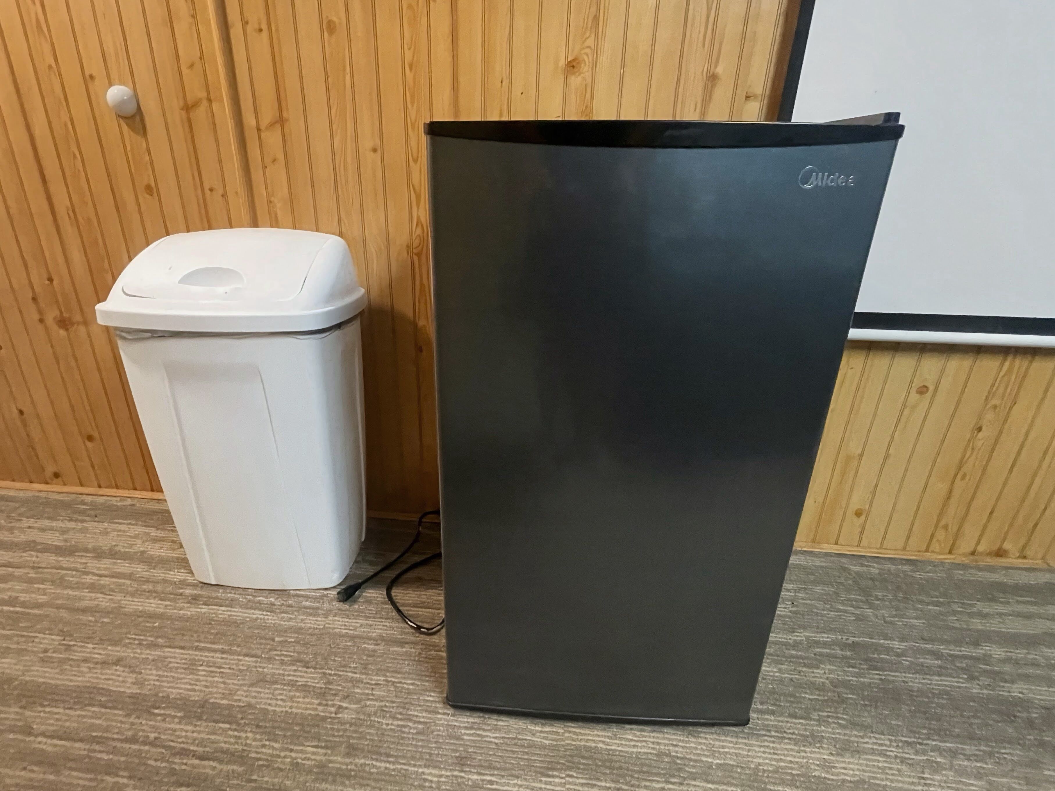 A white trash bin and a black appliance, possibly a refrigerator, next to a wooden wall with a whiteboard.