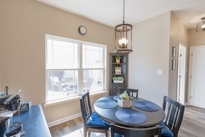 A dining area with a round table, blue placemats, and chairs. There is a hanging light fixture above the table.
