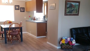 A dining area with wooden chairs and a table, a kitchen with wooden cabinets, a rug with animal prints, and a painting on the wall.