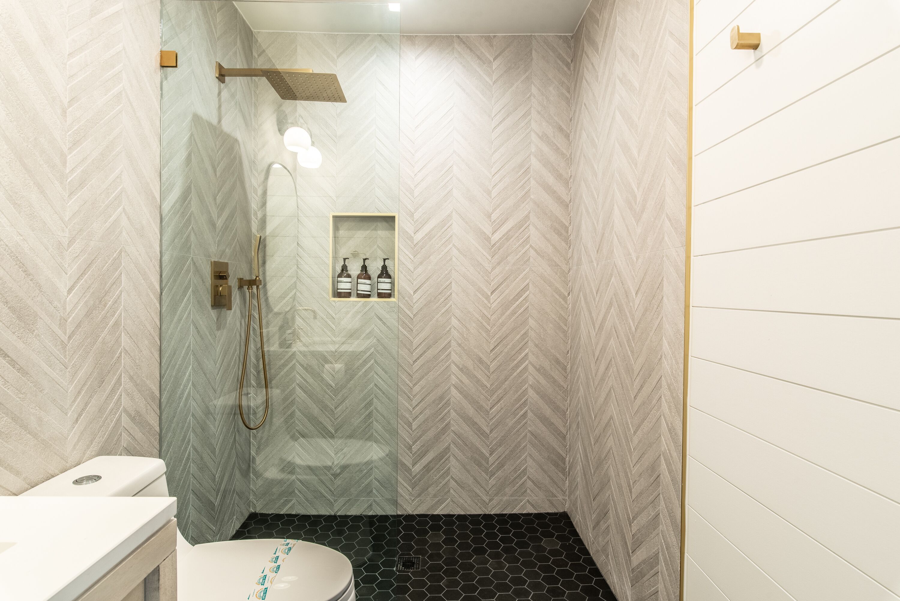 Un baño moderno con pared de diseño en patrón de espiga, ducha con cerramiento de vidrio y un inodoro con asiento blanco.