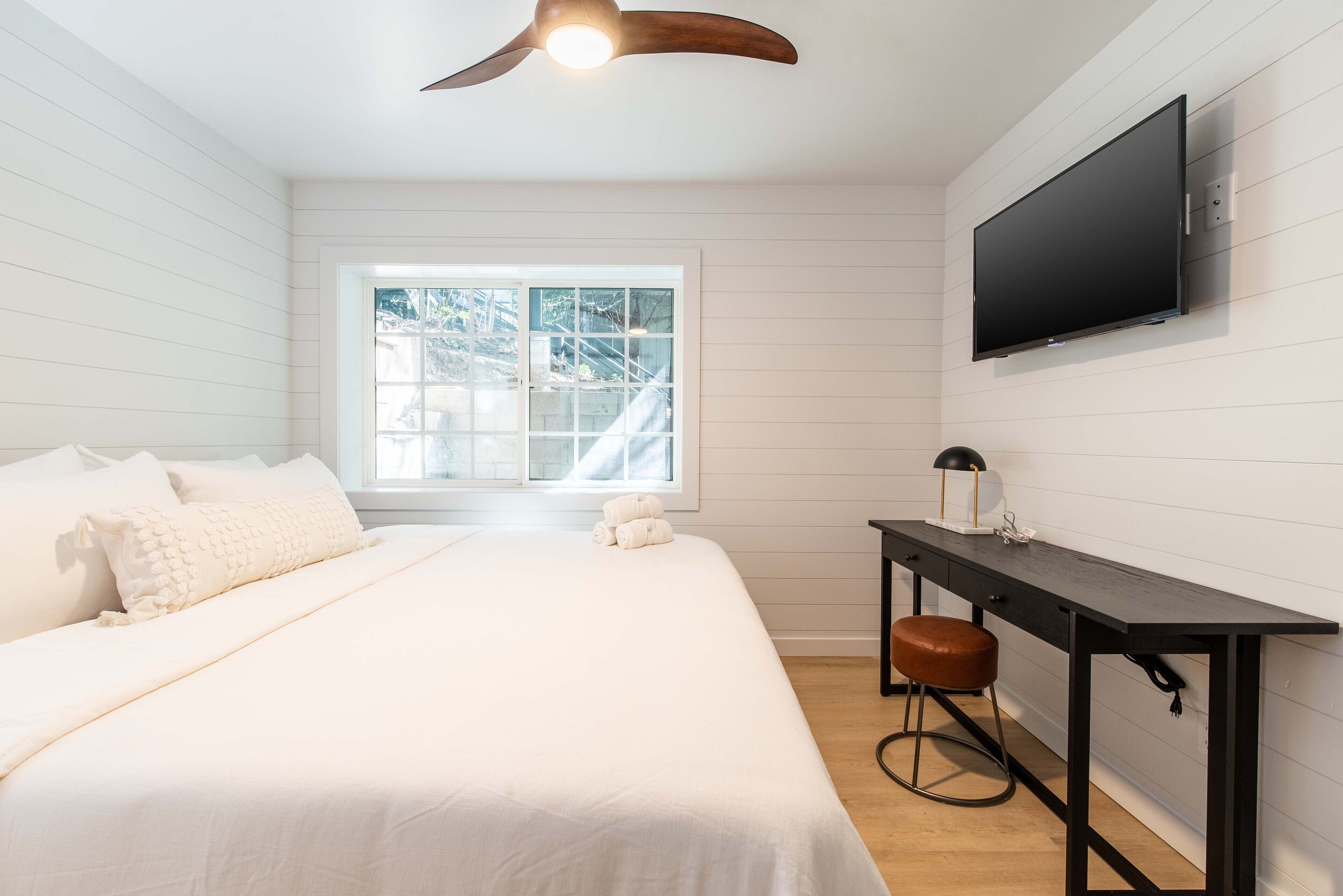 Un dormitorio con cama, escritorio, silla, televisor y ventana con vista a árboles.