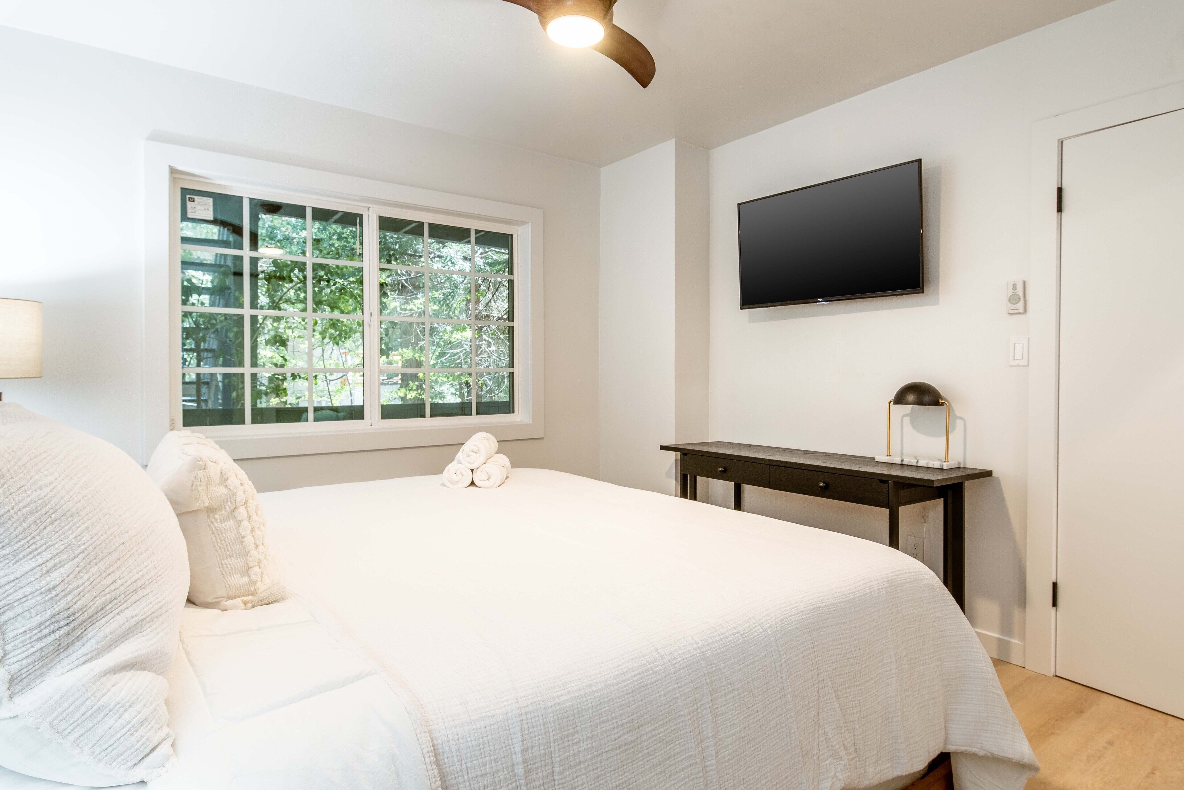 Quarto com cama grande, escrivaninha, TV de tela plana e janela com vista para árvores.