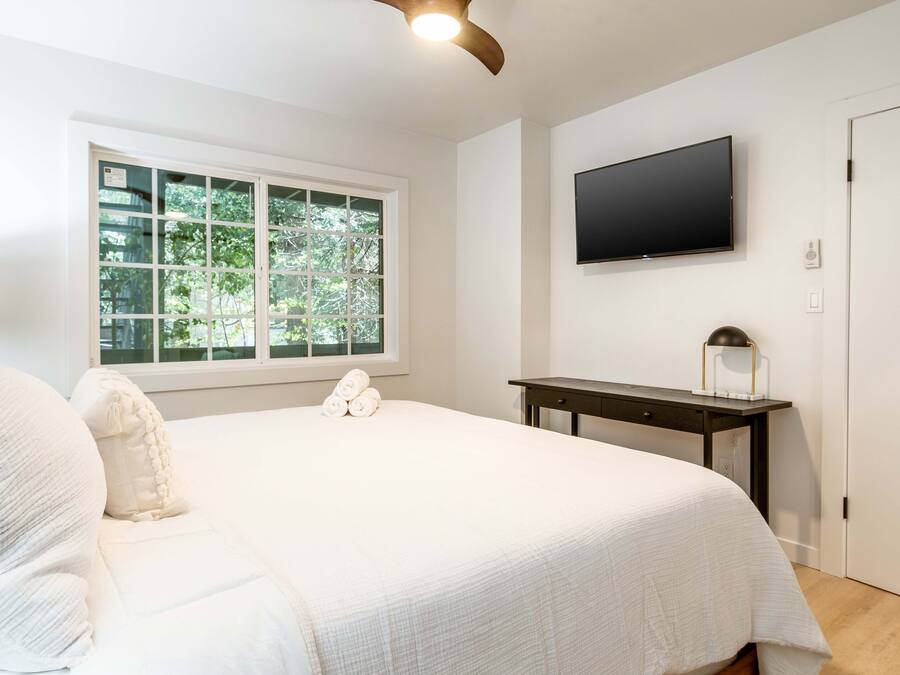 Quarto com cama grande, escrivaninha, TV de tela plana e janela com vista para árvores.