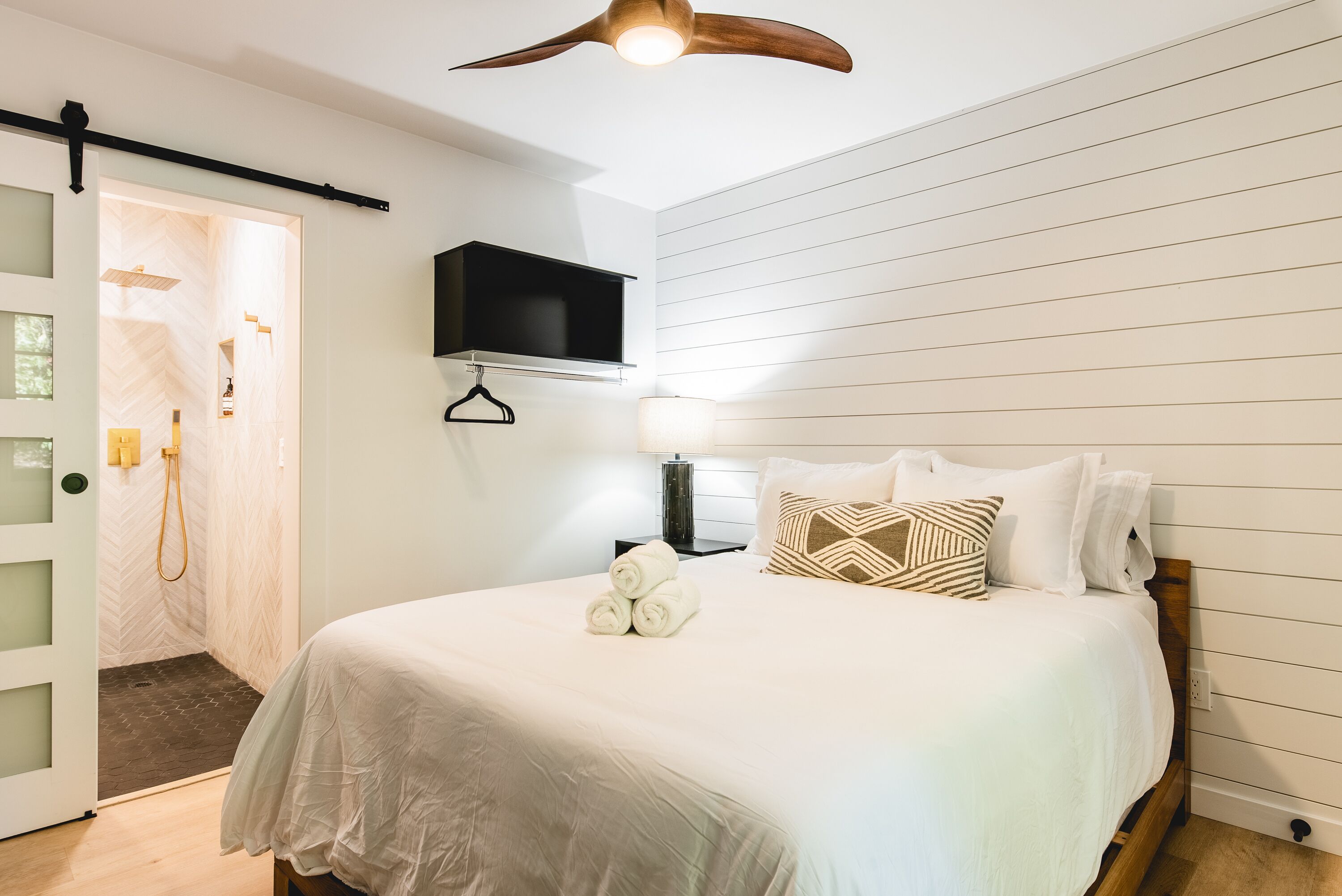 A bedroom with a bed, a TV mounted on the wall, a bedside lamp, and a wooden ceiling fan.