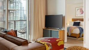 A modern hotel room with a large window, a flat-screen TV, a leather sofa, a bed with a colorful blanket, and a wooden floor.