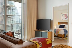Una habitación de hotel moderna con un ventanal grande, un televisor de pantalla plana, un sofá de cuero, una cama con un edredón de colores y piso de madera.
